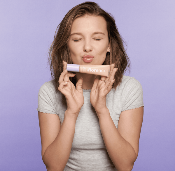 Millie Bobby Brown making a kissy face and holding foundation in front of a purple background